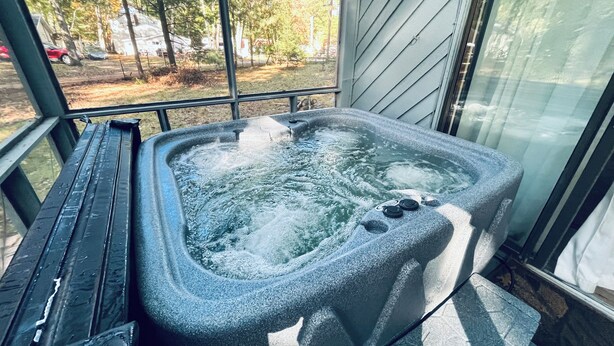 Un bain à remous à l’extérieur, avec un revêtement en pierre, placé près d’une fenêtre donnant sur des arbres et une voiture garée à l’extérieur.