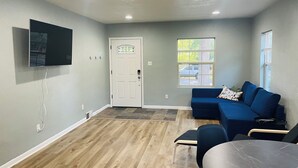 A room with a wooden floor, a white door with a stained glass window, a blue sofa, a small table, and a flat-screen TV mounted on the wall. - Cozy 3-bedroom house in charming Casper with AC, WiFi. Centrally located!  (Casper)