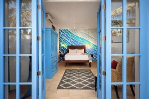 Ein Schlafzimmer mit einem Bett, einem Nachttisch, einem Korbstuhl, einem gemusterten Teppich und einer blauen Tür.