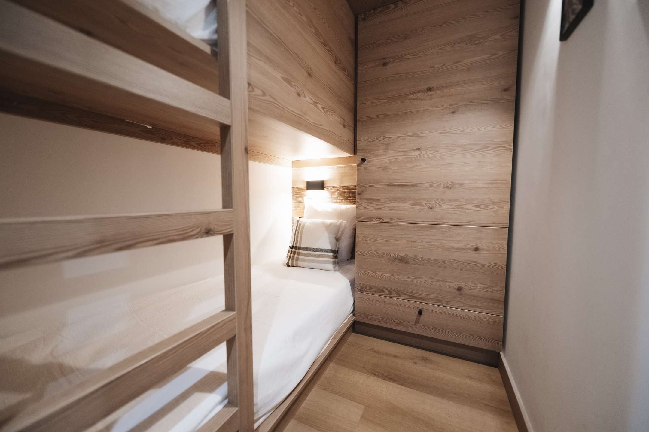 A narrow room with a bunk bed, a bedside table, and a wall-mounted light.