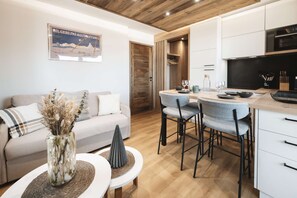 Een moderne woonkamer met een bank, salontafel en decoratieve vaas. Er is een eethoek met stoelen en een tafel gedekt voor twee personen. Een keuken met witte kastjes en een zwart aanrecht is zichtbaar.