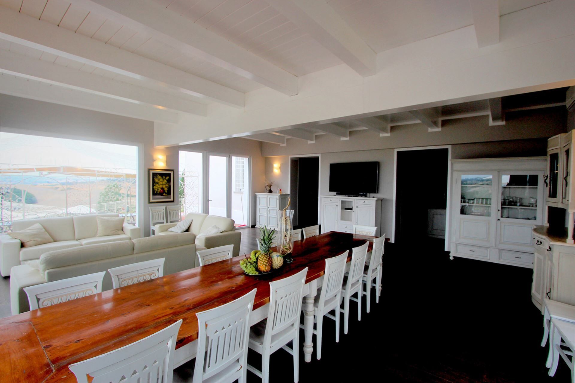 A spacious dining area with a long wooden table and white chairs, a white sofa, and a television mounted on the wall.