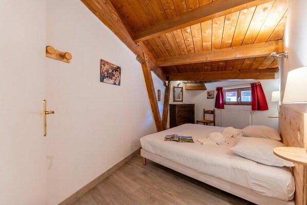 A bedroom with a wooden ceiling, a bed with white linens, a bedside table, and a window with red curtains.
