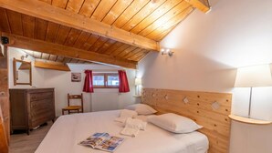 A bedroom with a wooden ceiling, a bed with white linens, a wooden dresser, a chair, a window with red curtains, and a bedside lamp. - Appartement Grand Roc 2, Argentière (Chamonix), France (Argentière (Chamonix))