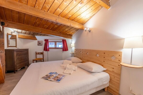 A bedroom with a wooden ceiling, a bed with white linens, a wooden dresser, a chair, a window with red curtains, and a bedside lamp. - Appartement Grand Roc 2, Argentière (Chamonix), France (Argentière (Chamonix))