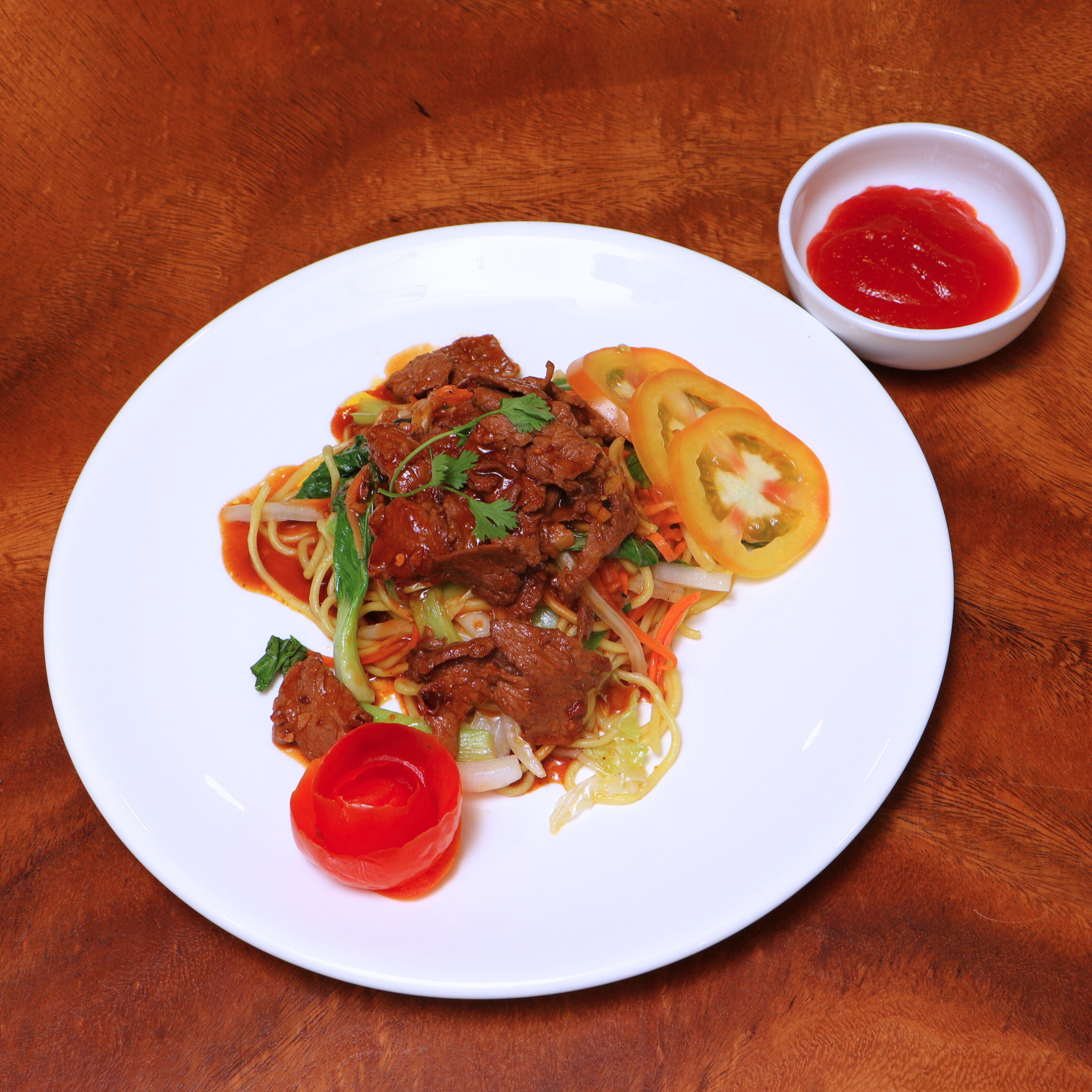 A plate of stir-fried noodles with beef, vegetables, and a small bowl of sauce on a wooden table.
