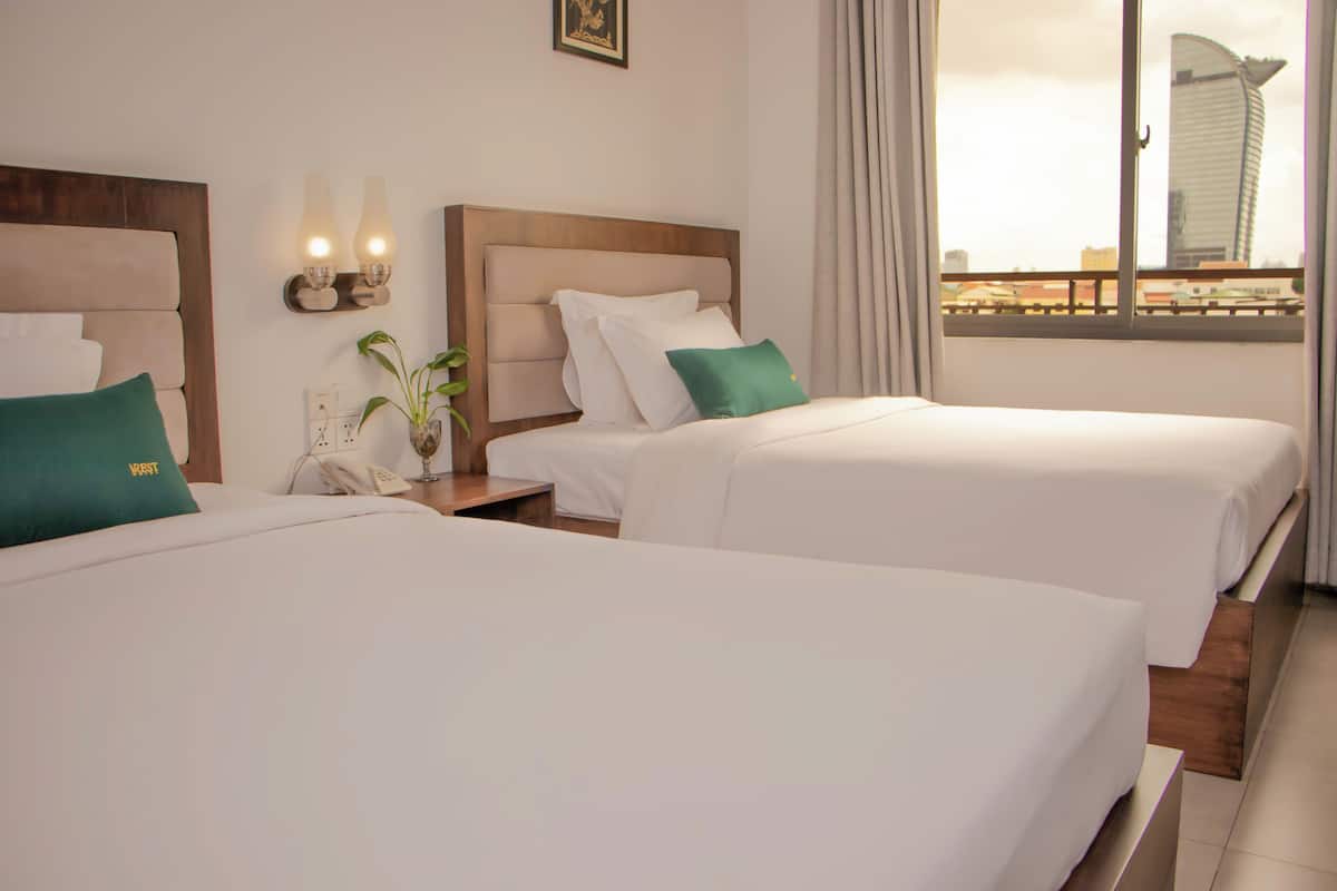 A hotel room with two beds, a nightstand with a phone and lamp, a window with a view of a building, and a plant on the nightstand.
