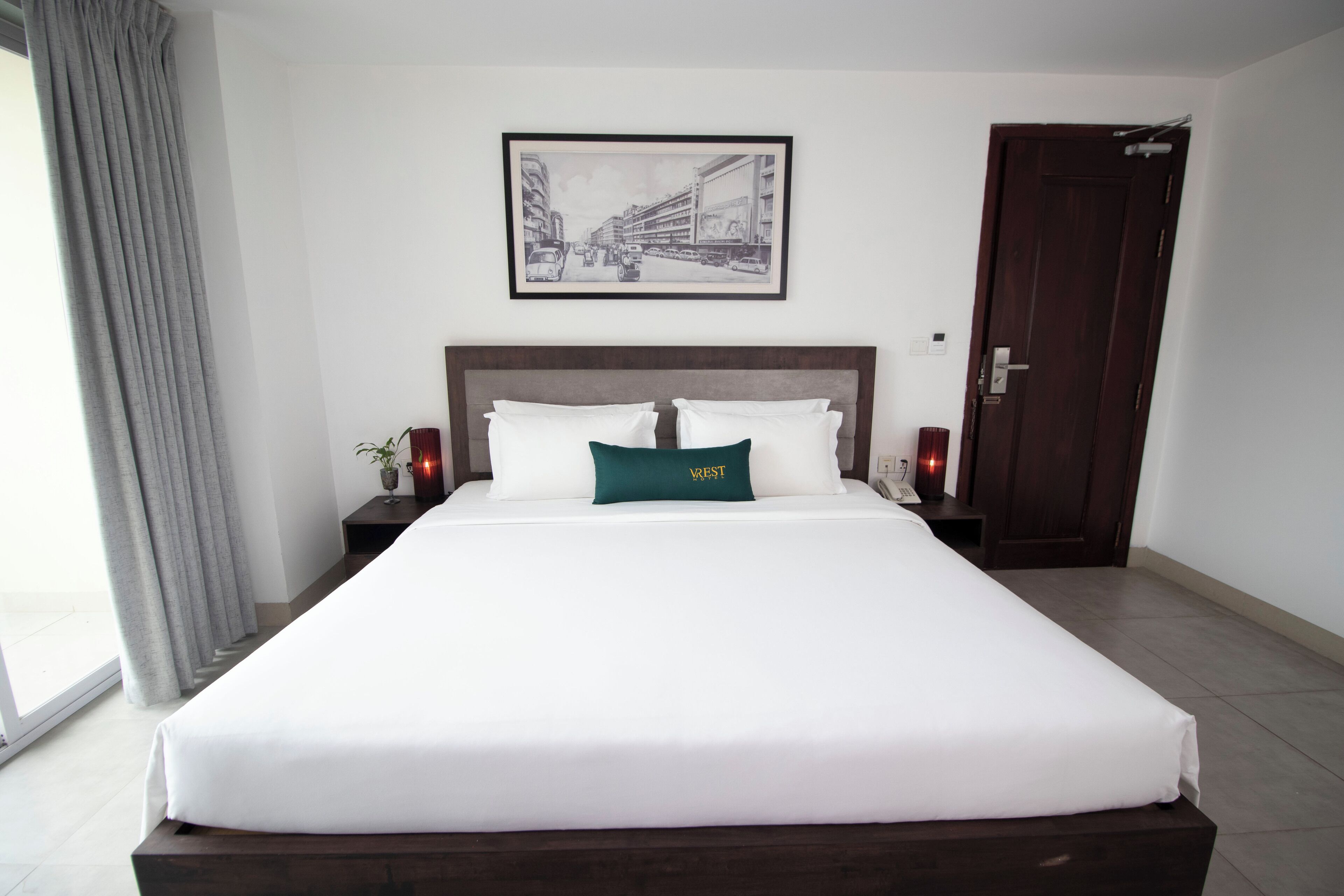 A large bed with white bedding and a green pillow, a framed picture on the wall, a dark wooden door, and a nightstand with a lamp.
