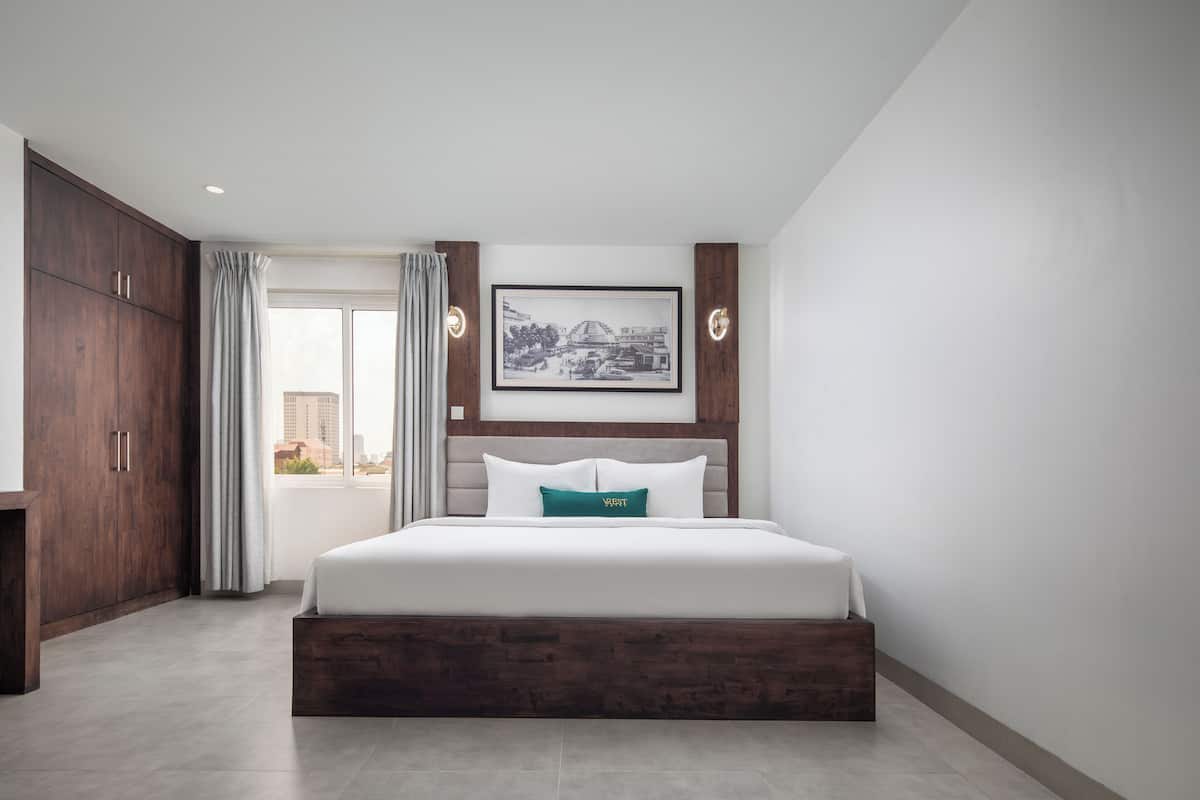 A bedroom with a large bed, a wooden wardrobe, a framed picture on the wall, and a view of buildings through a window.