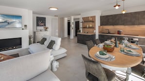 Un salon moderne avec une cheminée, un coin repas avec une table ronde et une cuisine dotée de meubles sombres et d’un éclairage suspendu.
