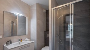 Une salle de bain moderne dotée d’une douche à parois de verre, d’un grand miroir et d’un meuble-lavabo blanc avec un évier et des tiroirs de rangement.