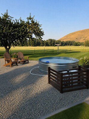 Un jacuzzi rodeado de grava, sillas de madera y un árbol en un entorno rural.