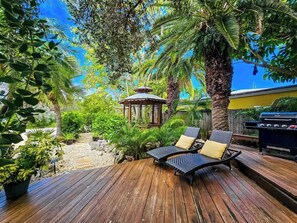 Um deck de madeira com duas cadeiras de descanso e um gazebo, tudo cercado por vegetação exuberante.