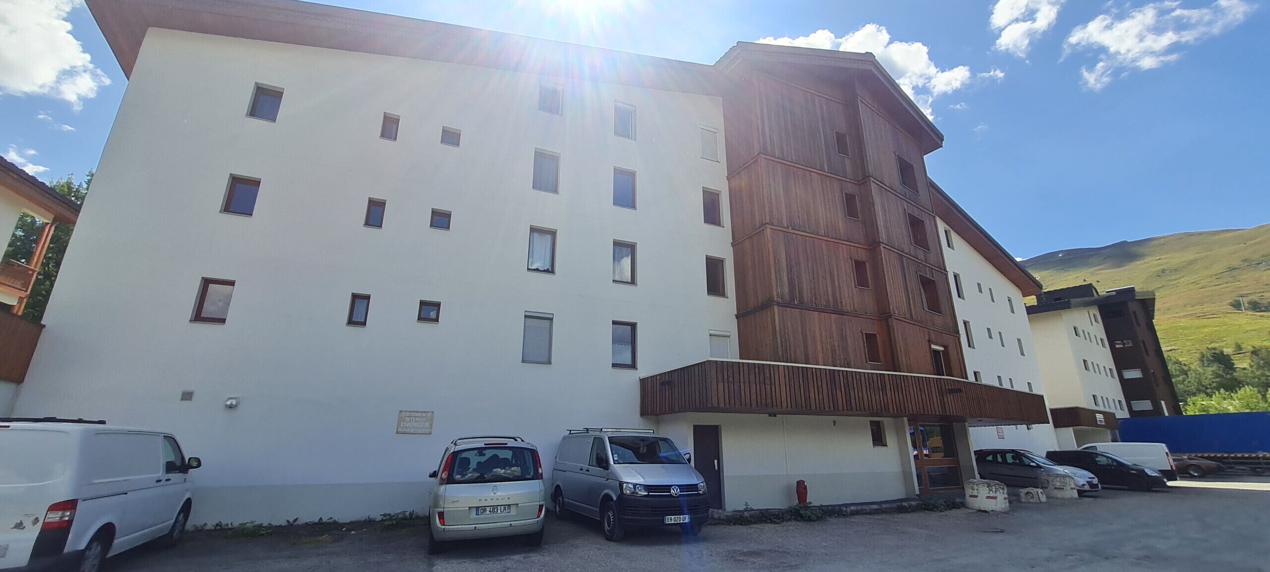 Un immeuble à plusieurs étages, doté d’une façade blanche et d’une partie en bois sur le côté droit. Plusieurs voitures sont garées devant.