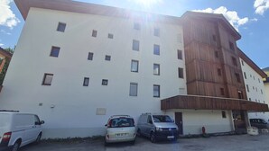 Ein mehrstöckiges Gebäude mit weißer Fassade und einem Holzbereich auf der rechten Seite. Vor dem Gebäude befinden sich mehrere parkende Autos.