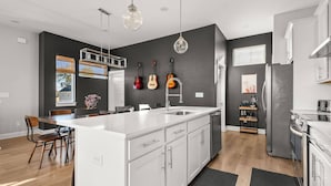 Cocina moderna con una isla grande, armarios blancos y salpicadero negro. Se observan dos guitarras colgadas, una estantería para vinos y una ventana con persianas.