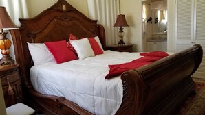 Una cama tradicional con cabecera de madera, ropa de cama blanca y detalles en rojo. Hay una mesita de noche con una lámpara, una silla y un tapete estampado en el suelo.