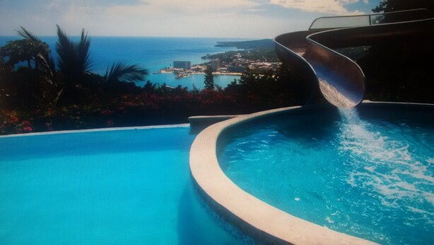Una piscina con borde curvo y un tobogán que desemboca en ella, rodeada de vegetación tropical y vistas a un pueblo costero.