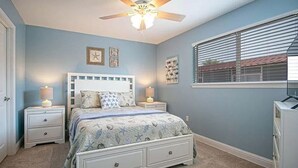 Une chambre à coucher avec un lit, des tables de chevet, un ventilateur de plafond et une fenêtre avec des stores.