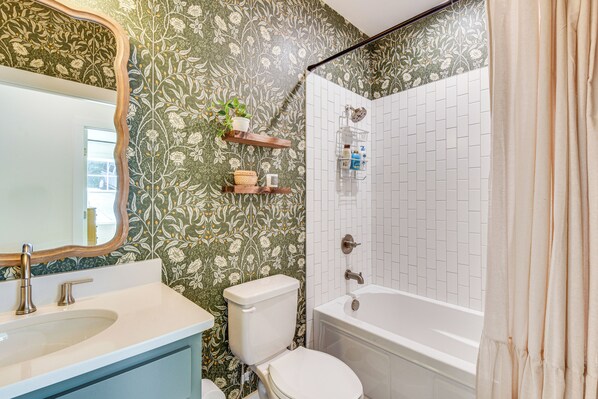 Casa de banho com sanita, lavatório e banheira. Existem prateleiras com produtos de higiene pessoal e um espelho.
