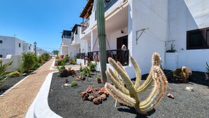 A courtyard with a variety of cacti and plants, a pathway, and a white building with balconies and windows. - Happy Beach Home - Shared pool By LVH (Puerto del Carmen)