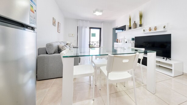 A modern living room with a glass dining table, white chairs, a grey sofa, and a flat-screen TV. - Happy Beach Home - Shared pool By LVH (Puerto del Carmen)