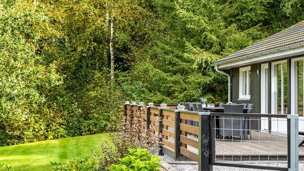 Terraza de madera con mobiliario exterior, rodeada por un jardín con diversas plantas y un muro de piedra.