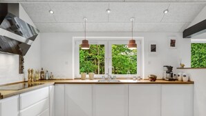 Una cocina moderna con armarios blancos, encimera de madera, pileta doble, luces colgantes y vistas a zonas verdes a través de amplios ventanales.