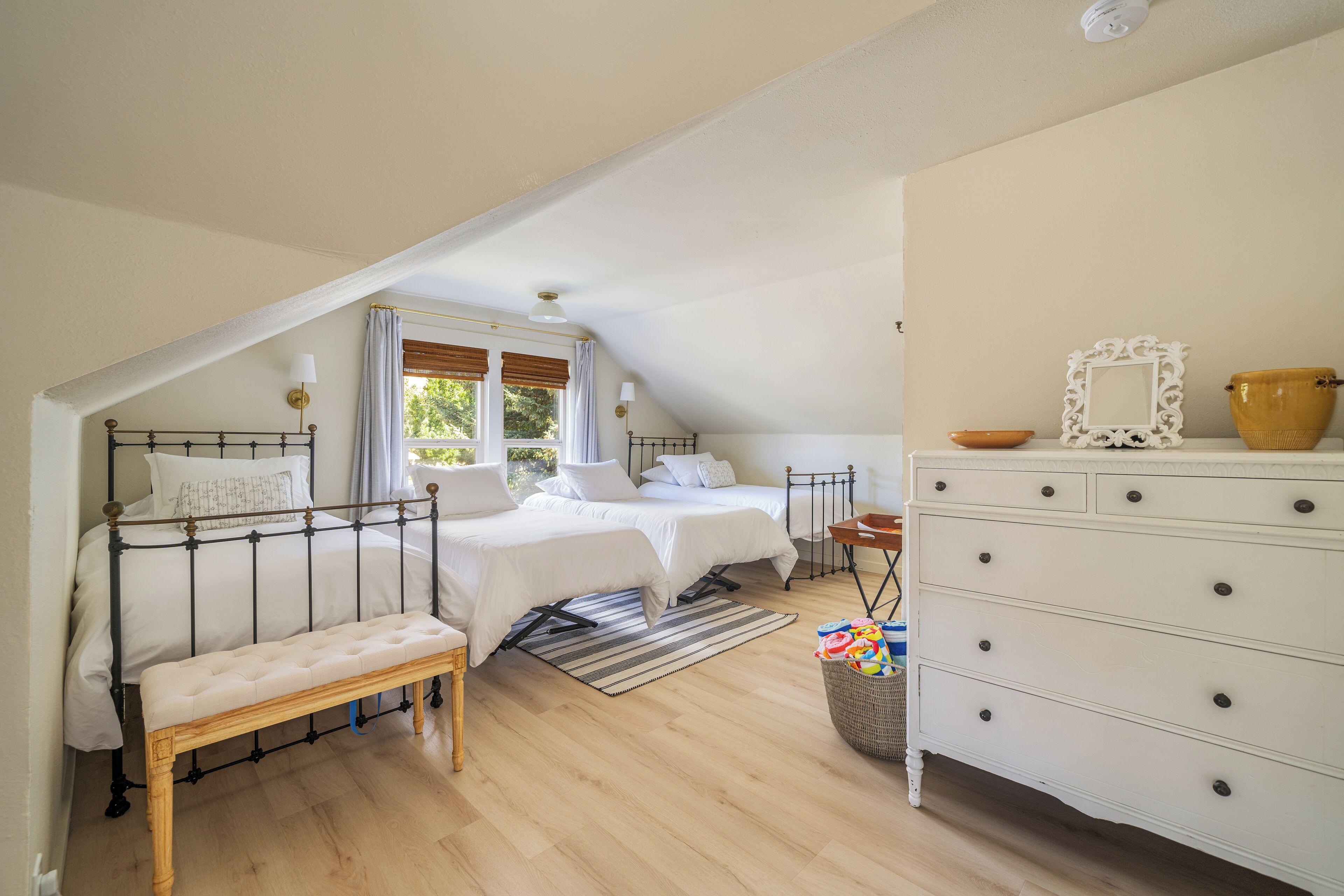 A bedroom with two beds, a bench, a dresser, and a window with curtains.