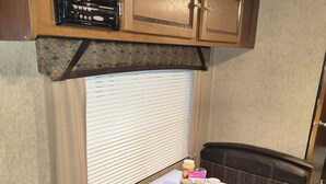 A compact kitchenette with a radio, a window with blinds, a small table with a tray of items, and a bench. - A well-maintained, cozy camper  in a secluded location near state land (Grayling)
