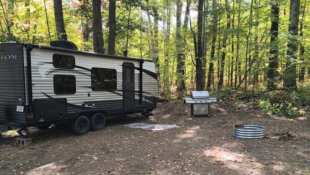 A recreational vehicle (RV) parked in a forested area with a portable grill and a metal container nearby. - A well-maintained, cozy camper  in a secluded location near state land (Grayling)