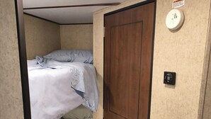 A compact bedroom with two beds, a wooden door, and a smoke detector on the wall. - A well-maintained, cozy camper  in a secluded location near state land (Grayling)
