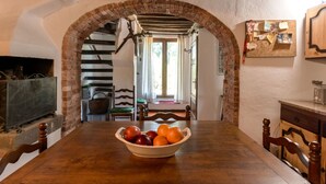 A rustic dining area with a wooden table, a bowl of fruit, and a brick archway leading to a kitchen.