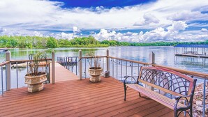 A wooden deck with a bench and potted plants, overlooking a calm lake with a dock and greenery in the background.