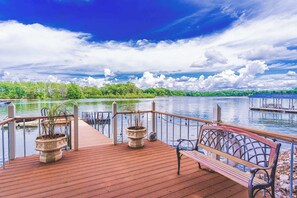 A wooden deck with a bench and potted plants, overlooking a calm lake with a dock and greenery in the background.