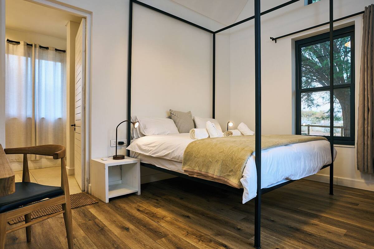 A bedroom with a four-poster bed, a nightstand, a chair, and a window with a view of trees.