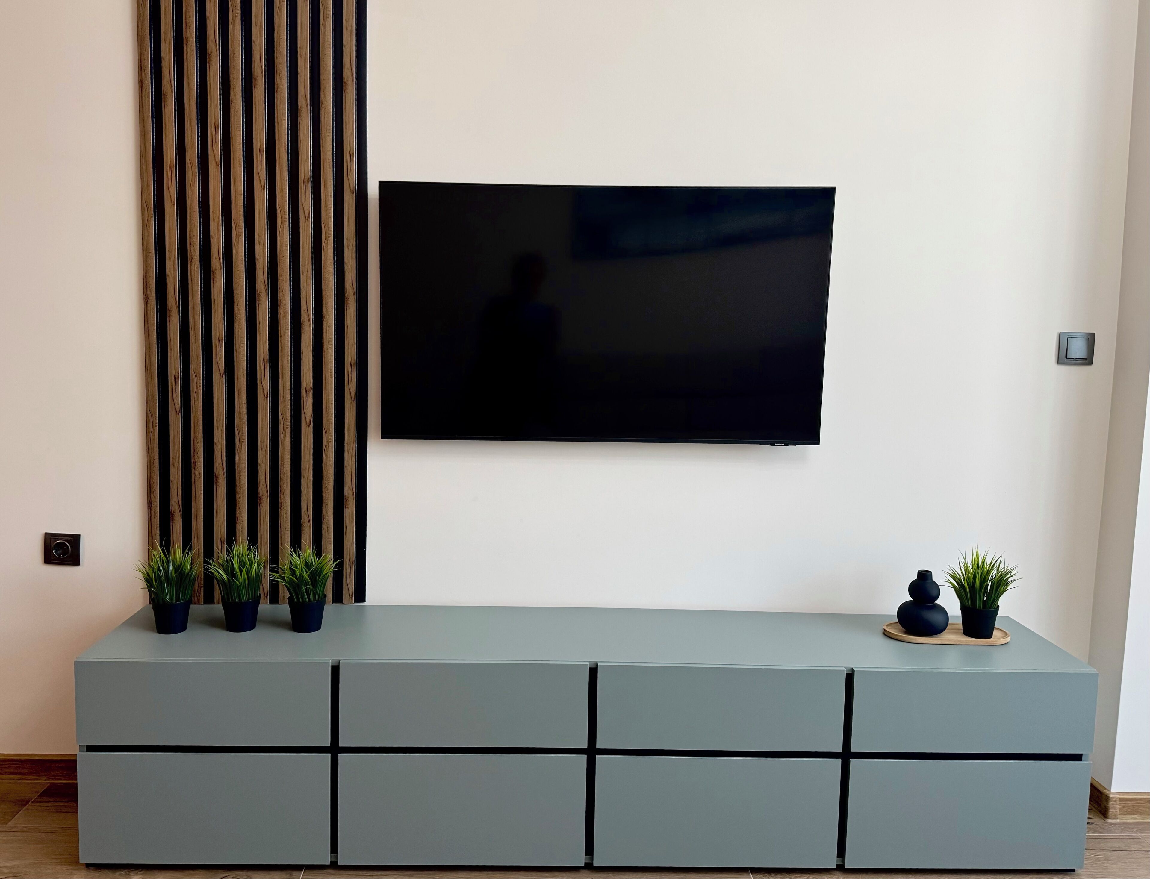 A modern living room with a flat-screen TV mounted on the wall, a grey entertainment unit with drawers, and two potted plants on the unit.