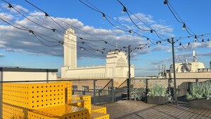 A rooftop terrace with yellow modular seating, a clear sky, and a large building with a tower in the background.