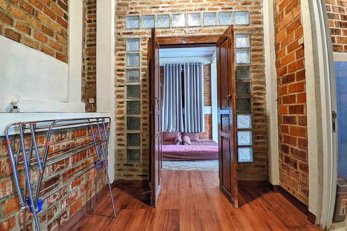 Um corredor estreito com piso de madeira, paredes de tijolo e uma porta que dá para um quarto com cama e cortinas.