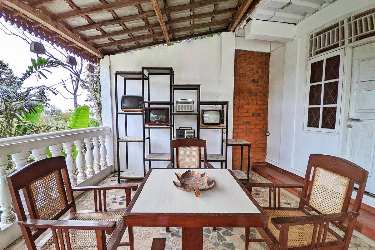 Uma área de estar ao ar livre coberta, com mesa e cadeiras, uma parede de tijolos e uma porta branca com janela.