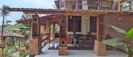 A rustic building with a covered patio, brick pillars, and a balcony overlooking a garden.