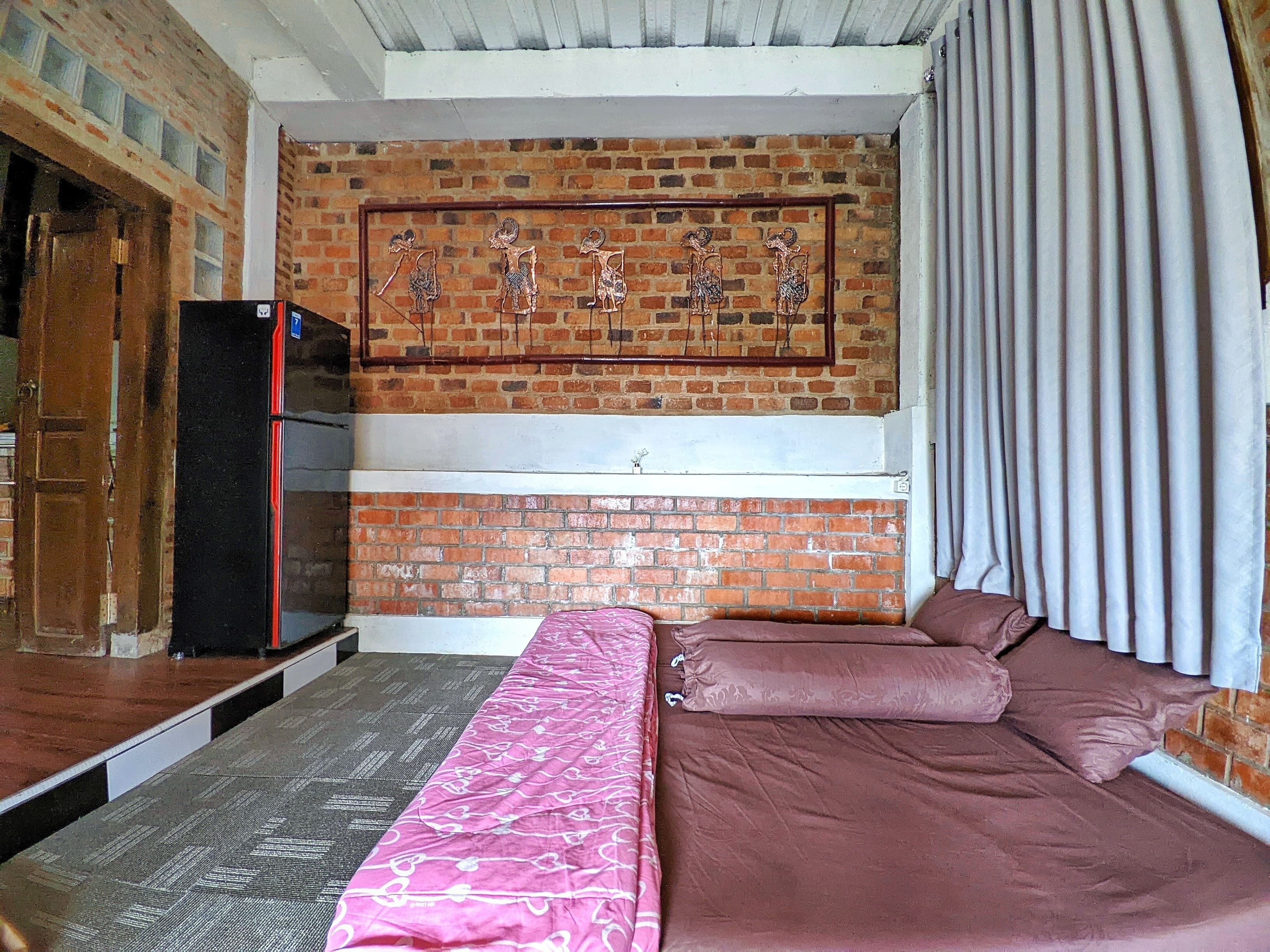 Een kamer met een bakstenen muur, een bed met roze en bruine beddengoed, een zwarte koelkast, en een raam met gordijnen.