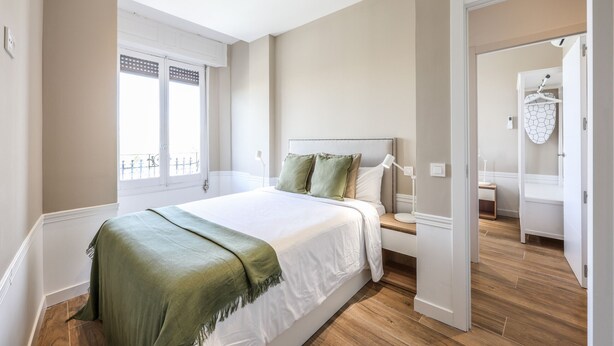 Un dormitorio con una cama grande, una mesita de noche, una ventana con persianas y una puerta que da a otra habitación.