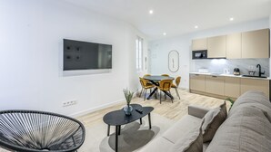 Una sala de estar moderna con un sofá, una silla negra, una mesa de comedor con sillas amarillas, una zona de cocina con armarios y un microondas, y un televisor de pantalla plana montado en la pared.