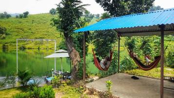A tranquil outdoor seating area with a swing chair and a hammock, overlooking a calm lake and lush greenery.