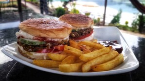 A plate with two sandwiches and a side of fries, set against a scenic view of a body of water and greenery. - Lake Wave Cottage & Resturent (Lekhnath)