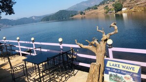 A lakeside dining area with tables and chairs, a sign for 'Lake Wave Cottage & Restaurant', and a view of the lake and surrounding hills. - Lake Wave Cottage & Resturent (Lekhnath)