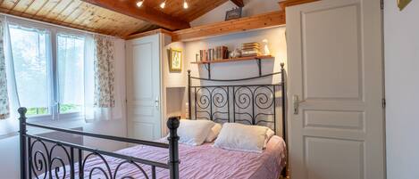 Une chambre à coucher avec un lit, des poutres en bois au plafond, une fenêtre avec des rideaux, une bibliothèque et une porte.