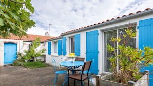 A courtyard with a round table and chairs, a small garden, and a building with blue shutters and a tiled roof. - Holiday Home 'Gîte Tout Confort 800m Plage' with Private Terrace and Private Garden (L'Épine)