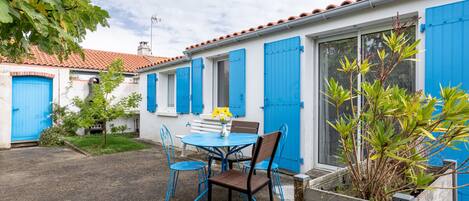 A courtyard with a round table and chairs, a small garden, and a building with blue shutters and a tiled roof.